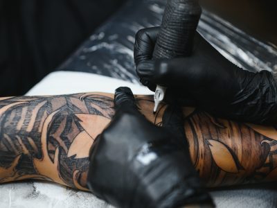 Cropped close up of a bearded tattoo artist working at his studio tattooing sleeve on the arm of his male client. Man getting tattooed by professional tattooist